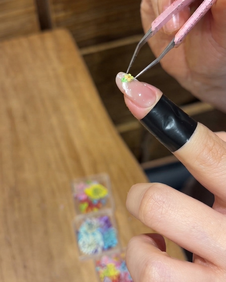 Pinzette di precisione che posizionano un minuscolo fiore secco giallo sulla superficie in gel bagnato di ununghia.
