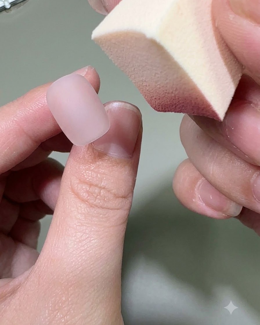 Una mano che tiene una spugnetta per trucco mentre applica il colore in gel rosa sulla punta di ununghia nude opaca.