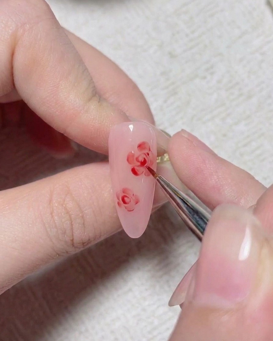 Un pennello fine per nail art aggiunge linee di dettaglio rosse per formare il centro di una rosa dipinta su ununghia rosa.