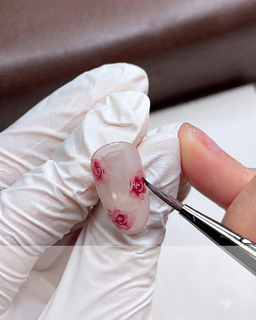 Una mano con i guanti che sfuma delicatamente i bordi di una rosa rosa ad acquerello su ununghia in gel usando un pennello fine.