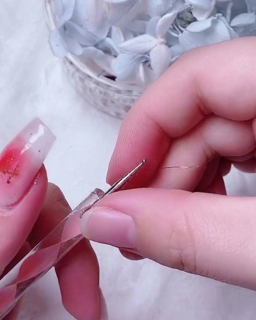 Pinzette di precisione che posizionano una piccola e intricata rosa in filo doro al centro di ununghia decorata.