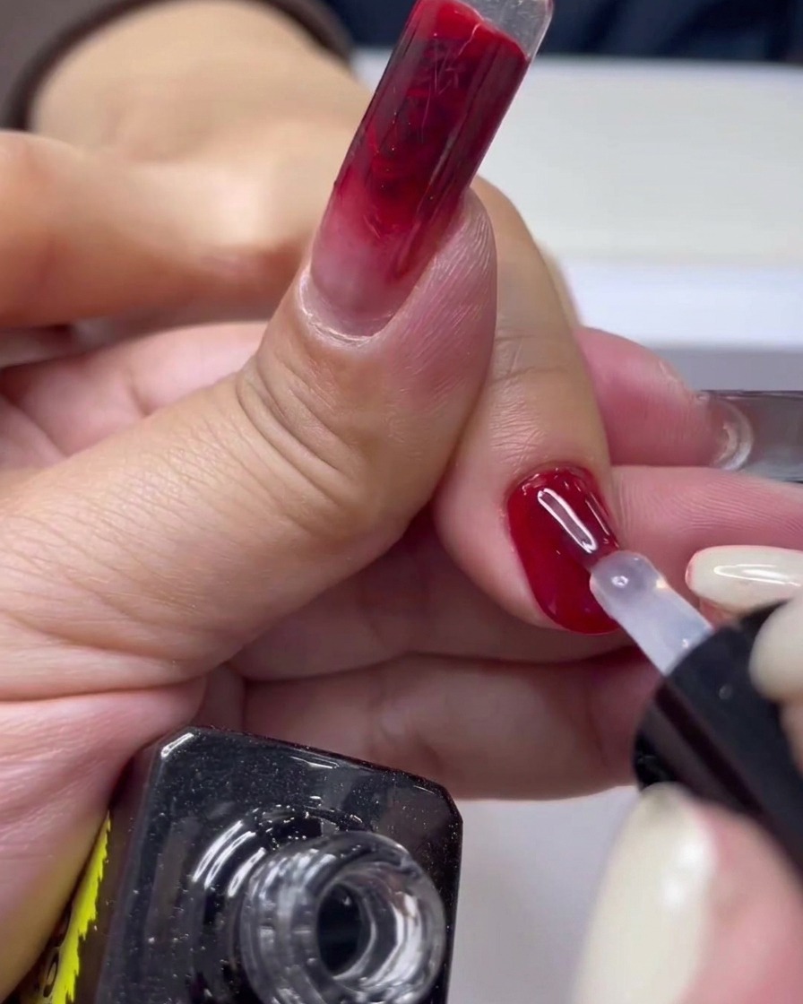 Un primo piano di un pennello da unghie che applica il top coat trasparente sopra una manicure gel rossa lucida.