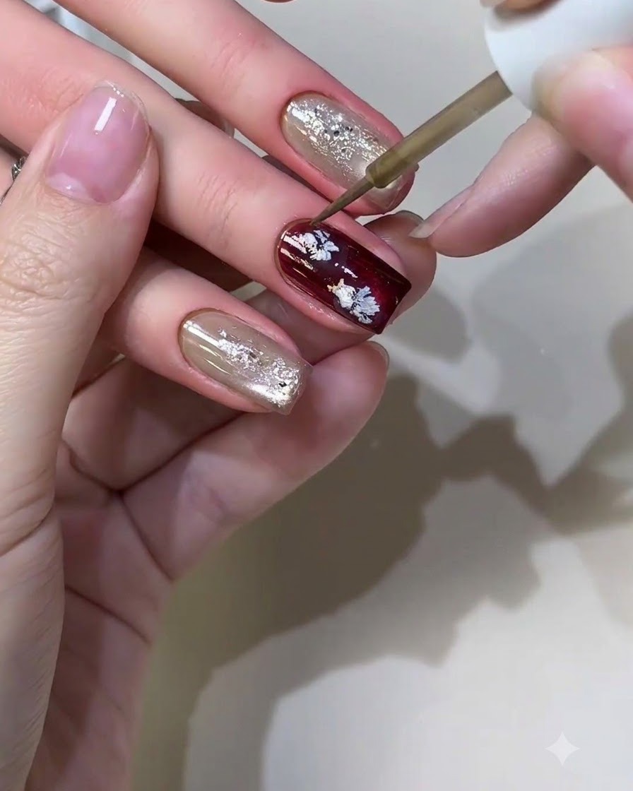 Un pennello fine per nail art che applica liquido metallico per formare piccoli motivi floreali casuali su ununghia borgogna.