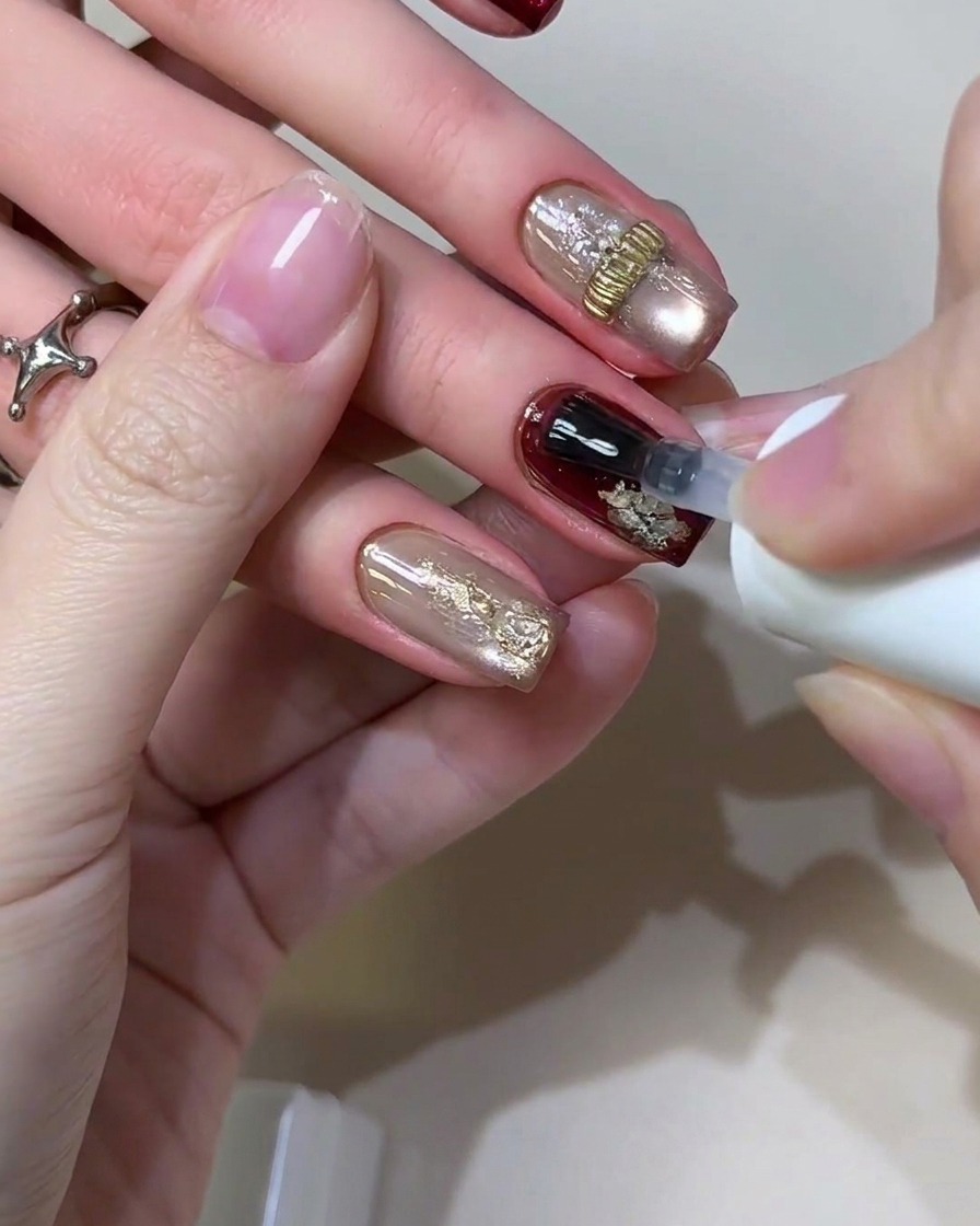 Un pennello per gel che applica un top coat sigillante trasparente e lucido su un design completato di unghie borgogna rétro e foglia doro.