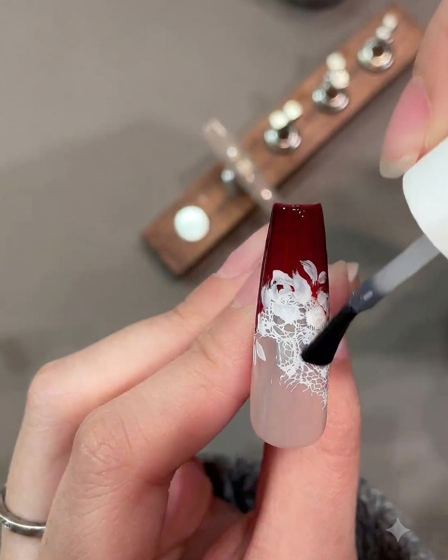 Un pennello applica un top coat opaco su un design per unghie in pizzo rosso scuro e bianco.