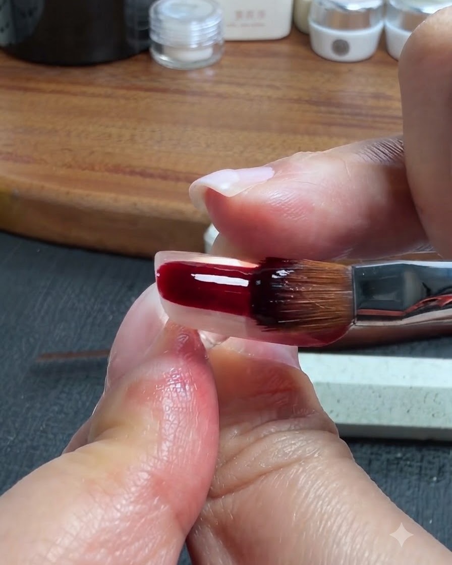 Un pennello per unghie che applica un gel traslucido rosso-marrone scuro sulla parte inferiore di ununghia artificiale trasparente.