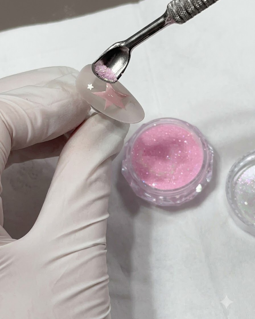 Un bastoncino spingi-cuticole in metallo che regge la polvere glitterata rosa sopra una stella dipinta su ununghia.