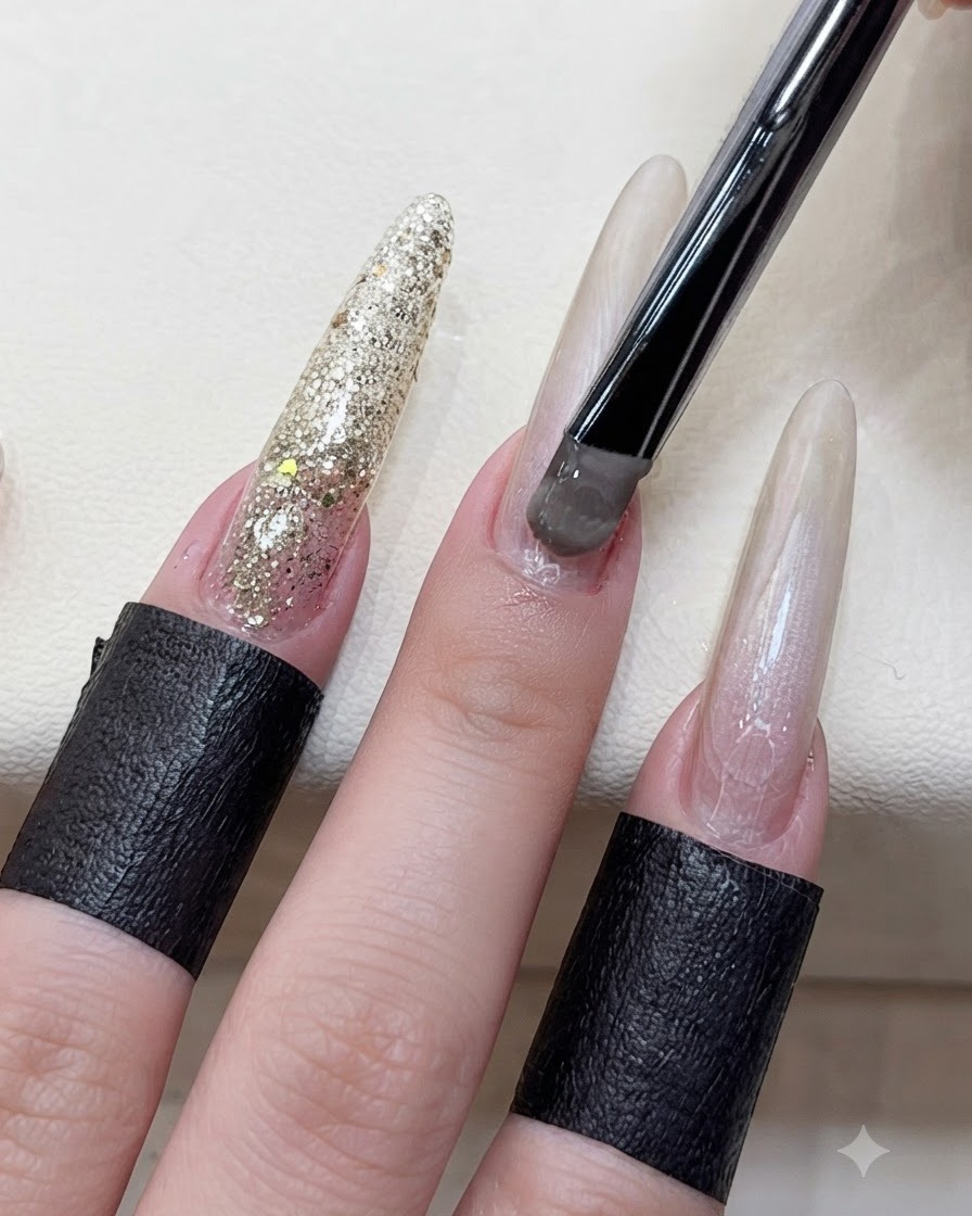 Primo piano di una mano che usa un pennello con manico nero per applicare il gel cat eye grigio-marrone sul letto ungueale di un dito allungato.