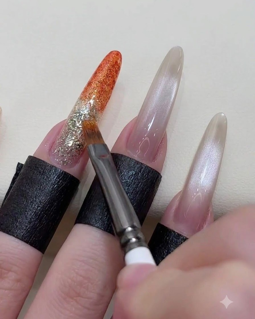 Primo piano di una mano che usa un pennello fine per sfumare e amalgamare la linea di transizione di un gel arancione vibrante applicato su una base glitterata.