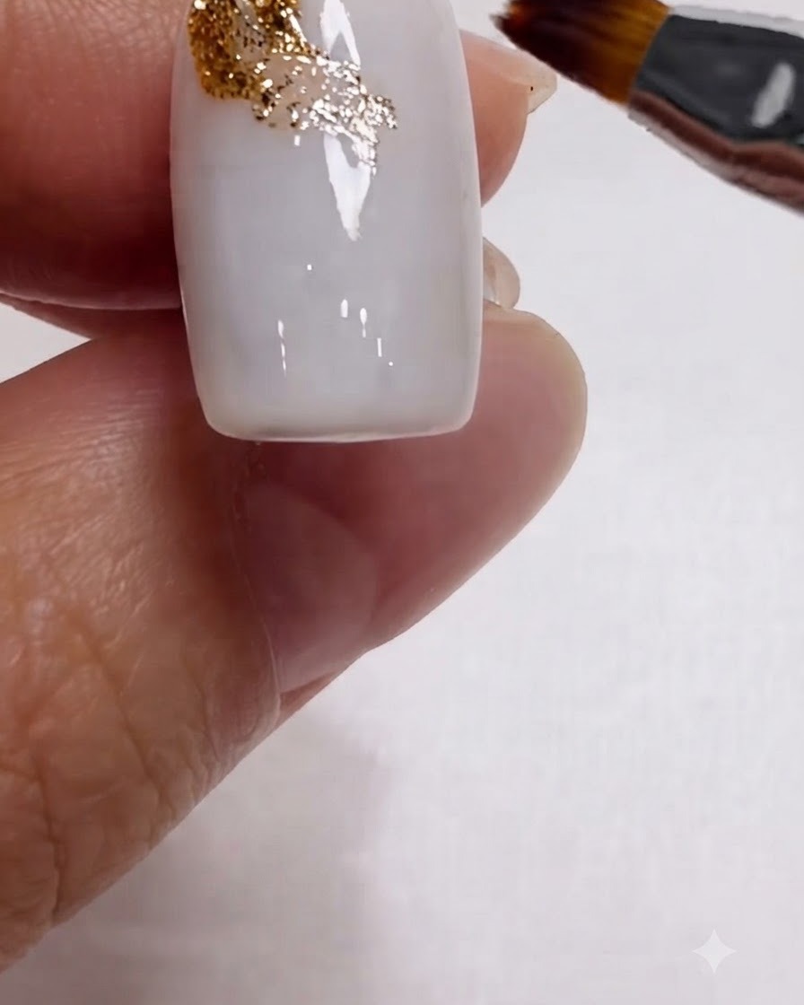 Un pennello che sfuma diagonalmente il glitter dorato sullangolo superiore di ununghia bianco latte.