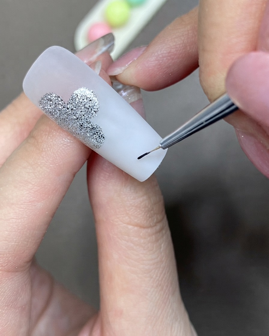 Un pennello per dettagli sottili che applica il gel liner nero per creare una forma a trifoglio accanto a un design in glitter argento.