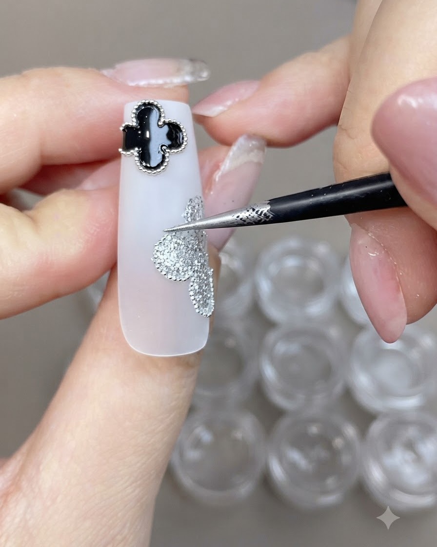 Un pennello sottile che posiziona piccole sfere in acciaio lungo il bordo di un design a trifoglio glitter argento su ununghia bianca.