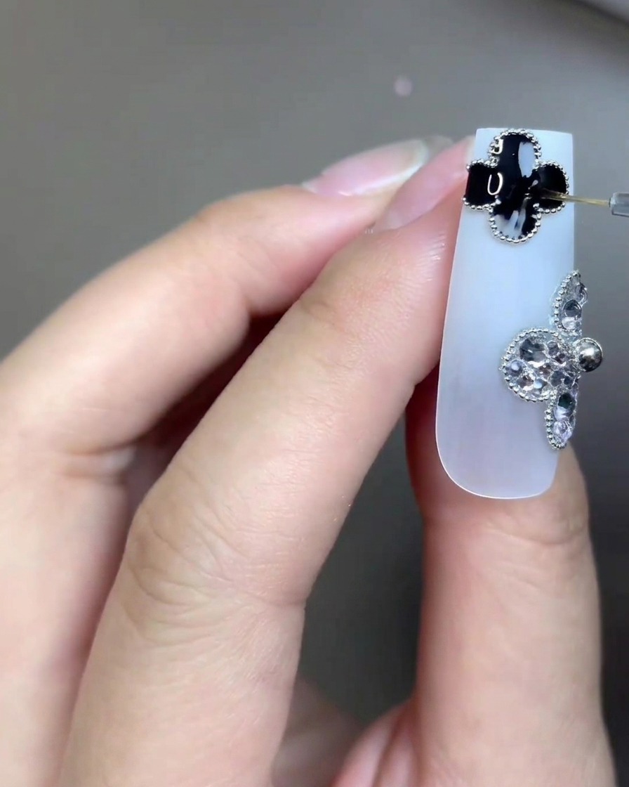 Un primo piano di ununghia lunga bianco latte decorata con due ciondoli a trifoglio a quattro foglie, uno nero e uno di strass, mentre un pennello per nail art molto fine applica il top coat trasparente sul dettaglio del trifoglio nero.
