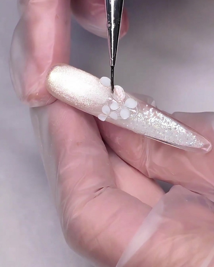 Un pennello da dettaglio fine che applica il gel cat-eye rosa nude su un fiore bianco scolpito in 3D su ununghia.