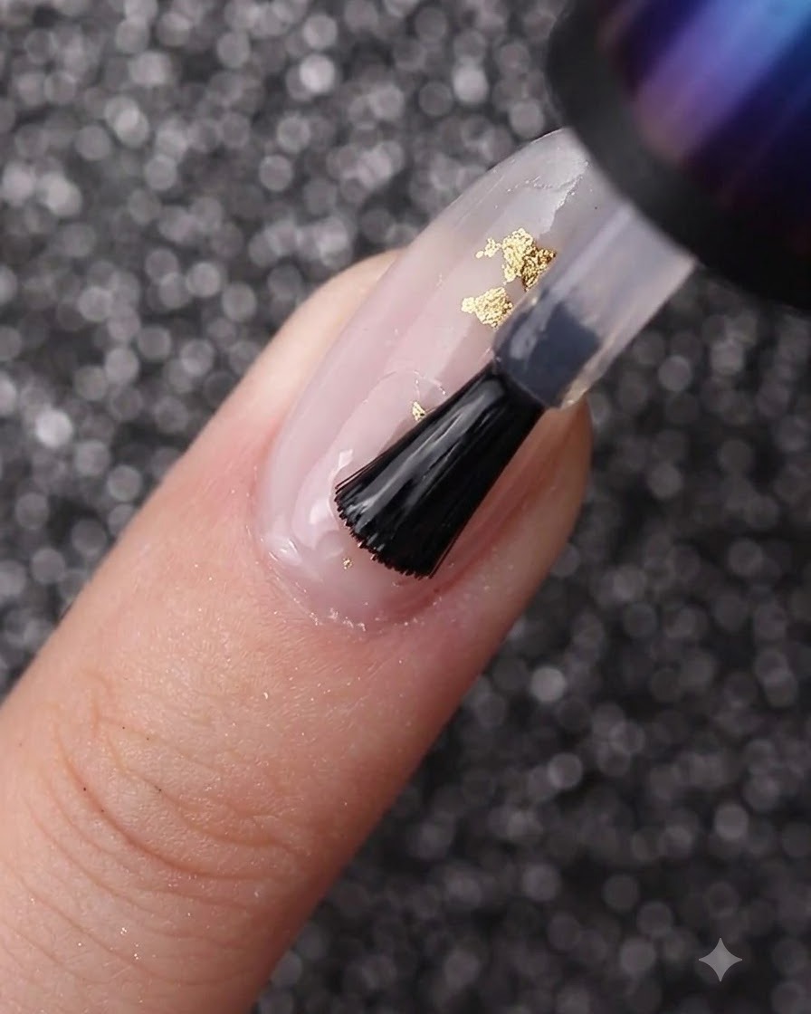 Un pennello da unghie che stende il gel trasparente sopra ununghia decorata con smalto bianco latte e foglia oro.