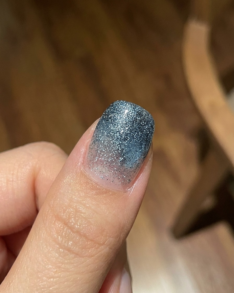 Un dito che mostra una mano di gradiente gel jelly blu traslucido su uno sfondo strutturato e glitterato, scattato in piena luce naturale.