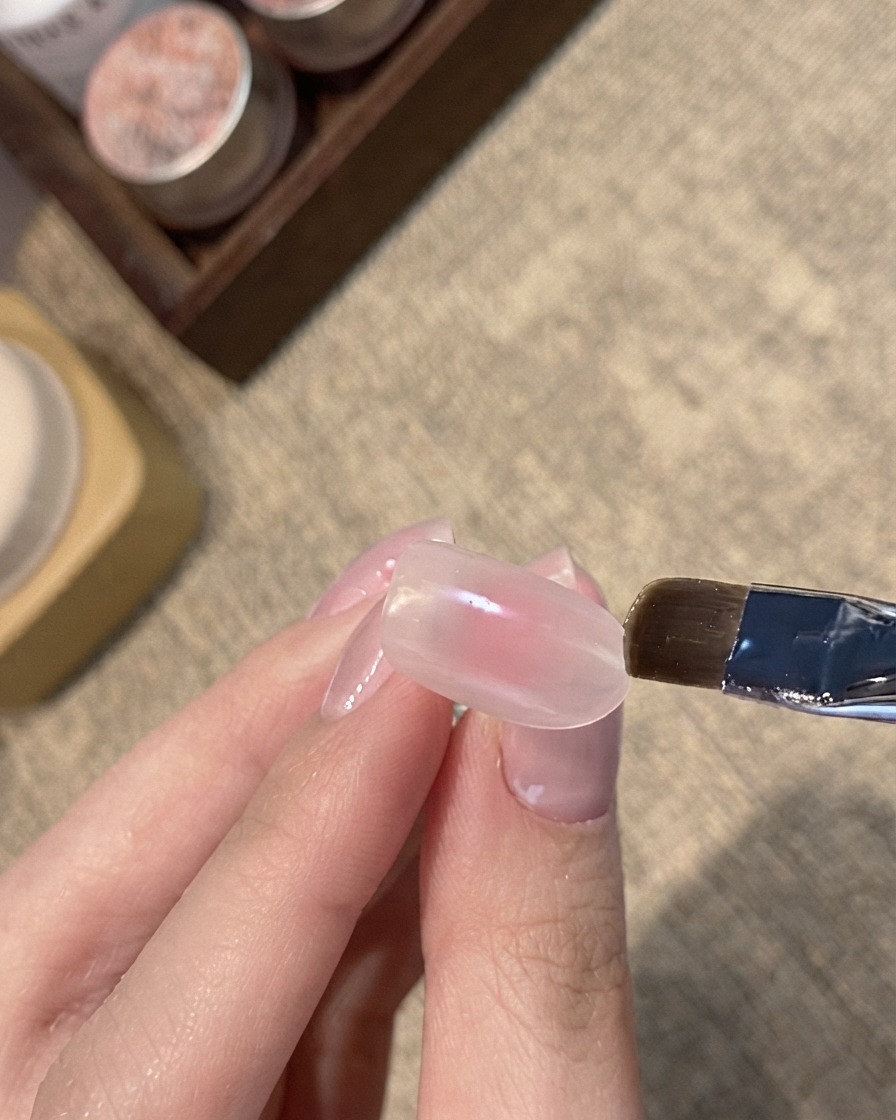 Un pennello piatto che applica un gel cat-eye shimmer blu sulla punta di ununghia rosa nude.