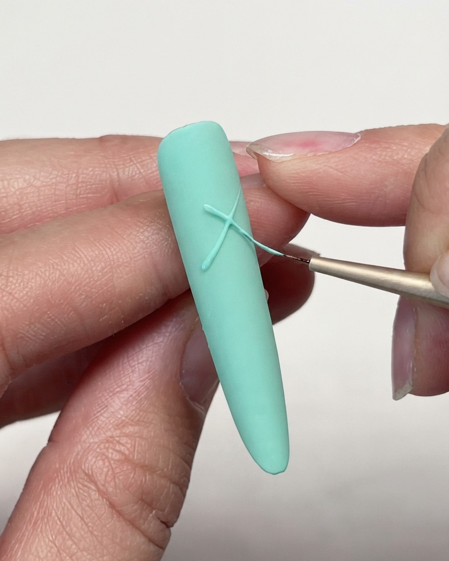 Primo piano di un pennello liner sottile che incrocia linee di gel su ununghia opaca verde menta per iniziare il design della stella.
