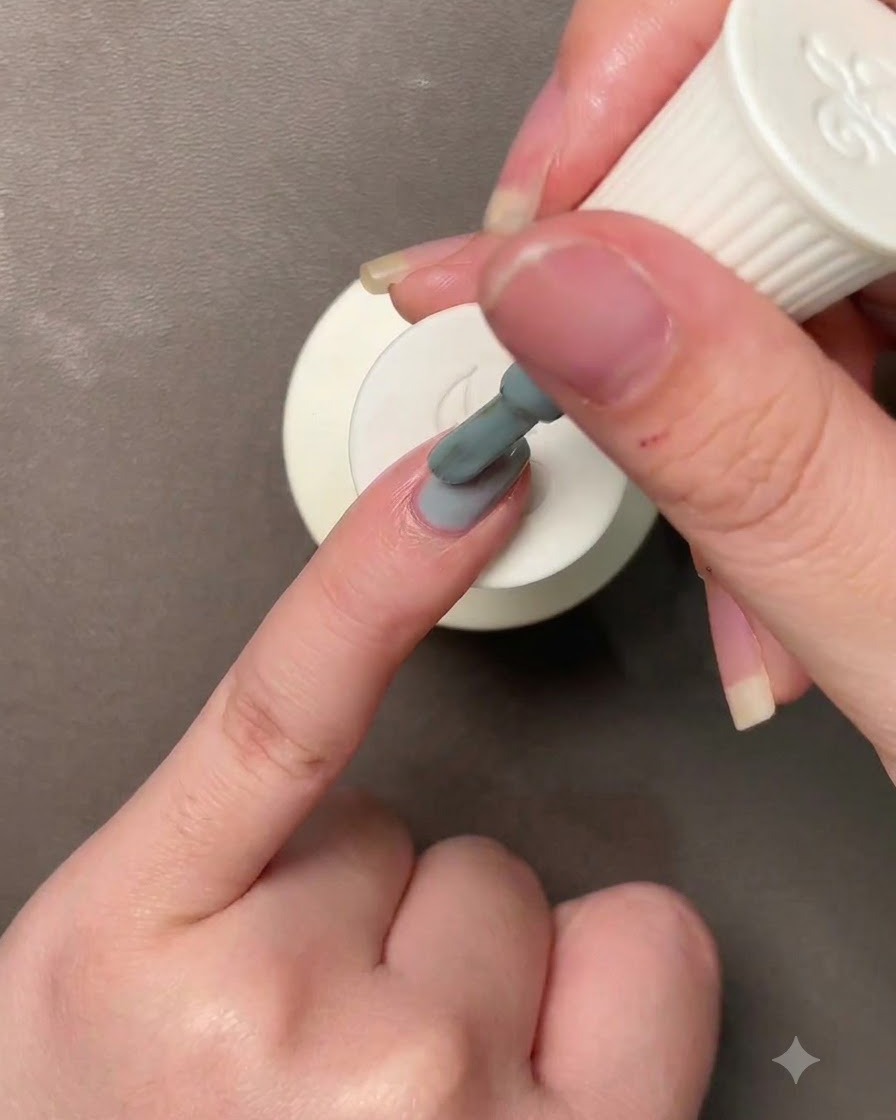 Una mano che tiene un pennello applicando uno smalto gel blu-verde Morandi su ununghia.