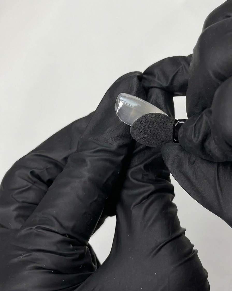 Una mano con guanto nero che usa un applicatore in spugna per strofinare polvere cromata iridescente su una tip di unghia finta liscia.