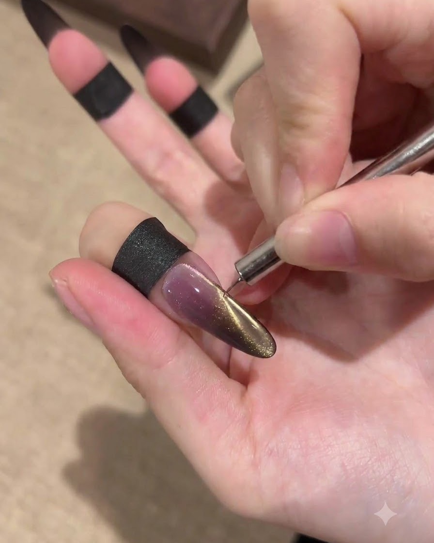 Una mano utilizza un sottile strumento magnetico vicino al bordo laterale di ununghia a stiletto lunga per attirare un effetto cat-eye dorato su una base scura.