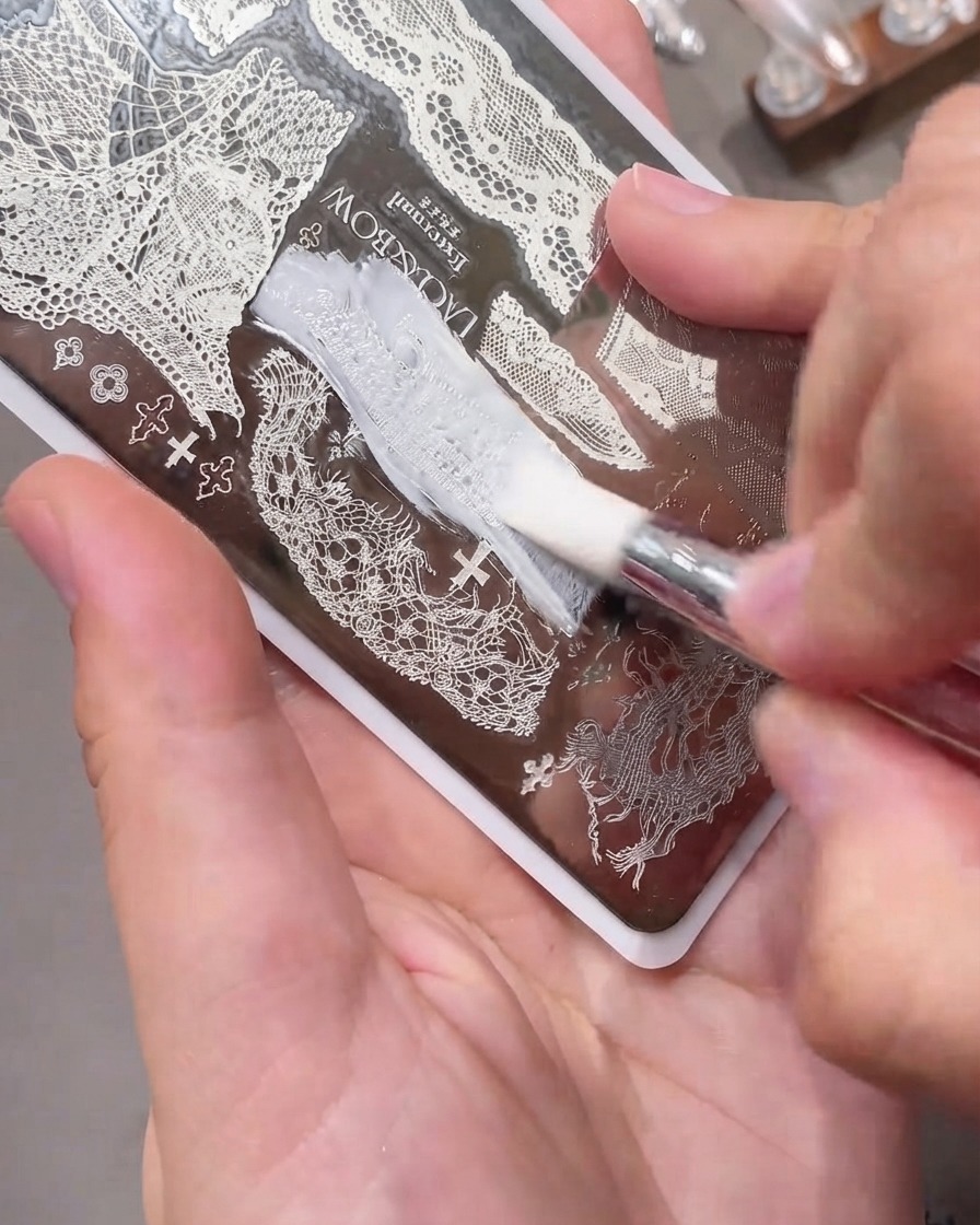 Una carta raschietto scivola dolcemente su una piastra per stamping in metallo incisa piena di smalto gel bianco.