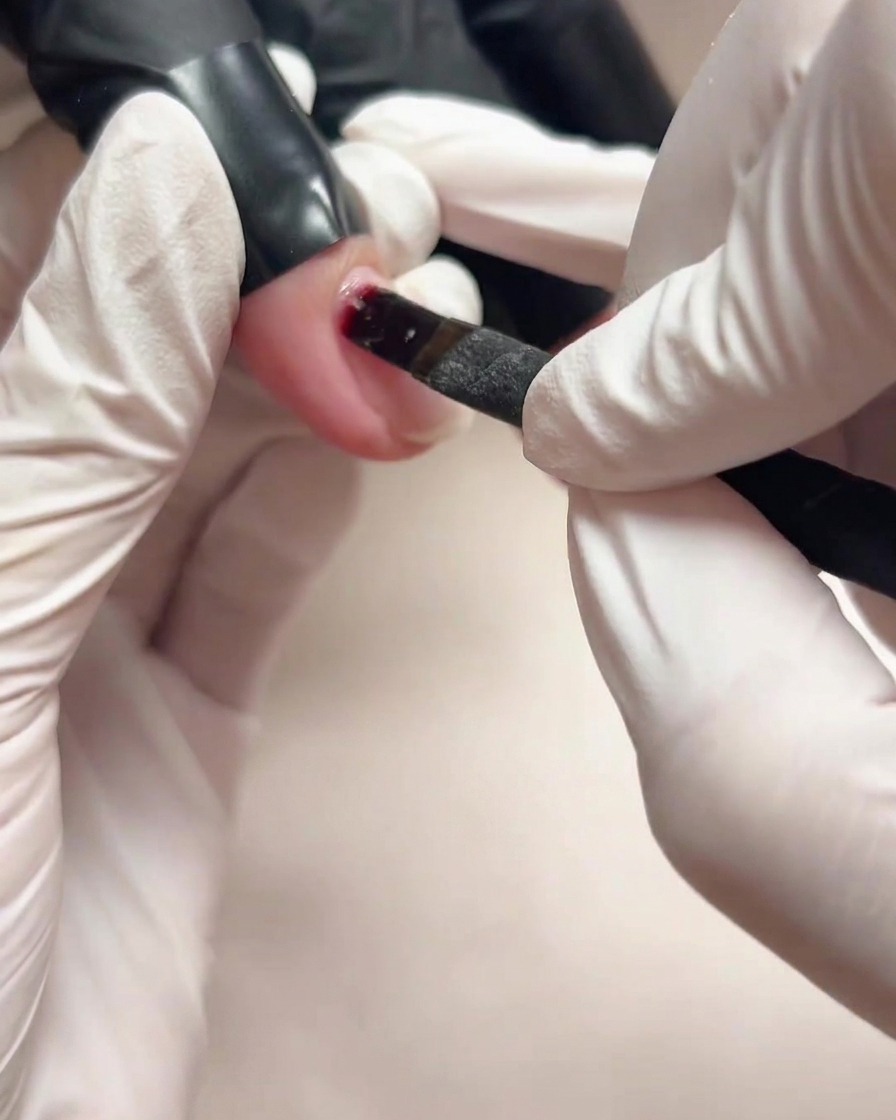 Primo piano di una mano con guanto che usa un piccolo pennello per applicare il gel base trasparente su ununghia naturale.