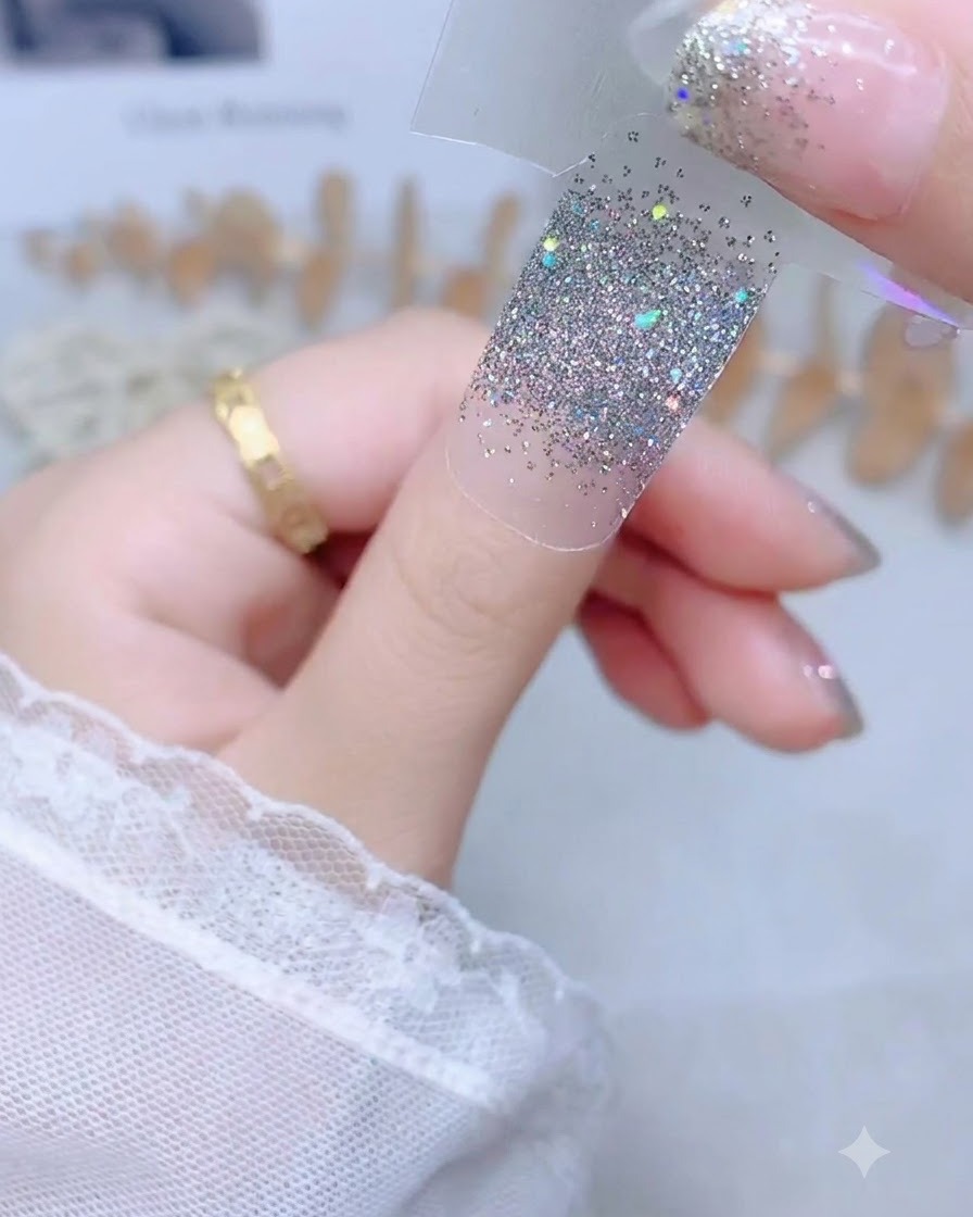 Una mano che tiene un adesivo per unghie glitter argento da una striscia di supporto trasparente sopra ununghia del pollice nuda.