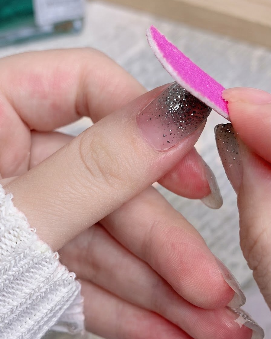 Una lima in cartone rosa che lima via la porzione in eccesso di uno sticker per unghie con glitter argento su un pollice.