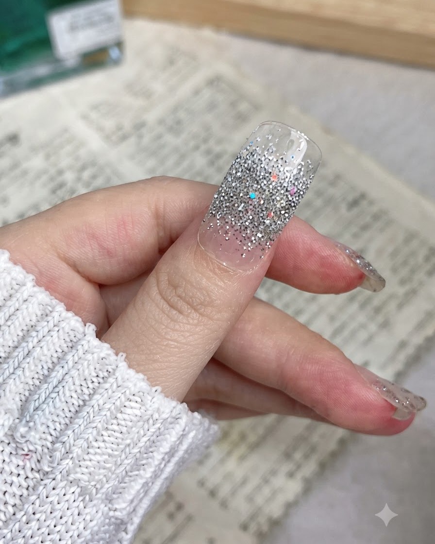 Un primo piano di un pollice con un adesivo per unghie glitter argento applicato uniformemente dalle cuticole al margine libero.