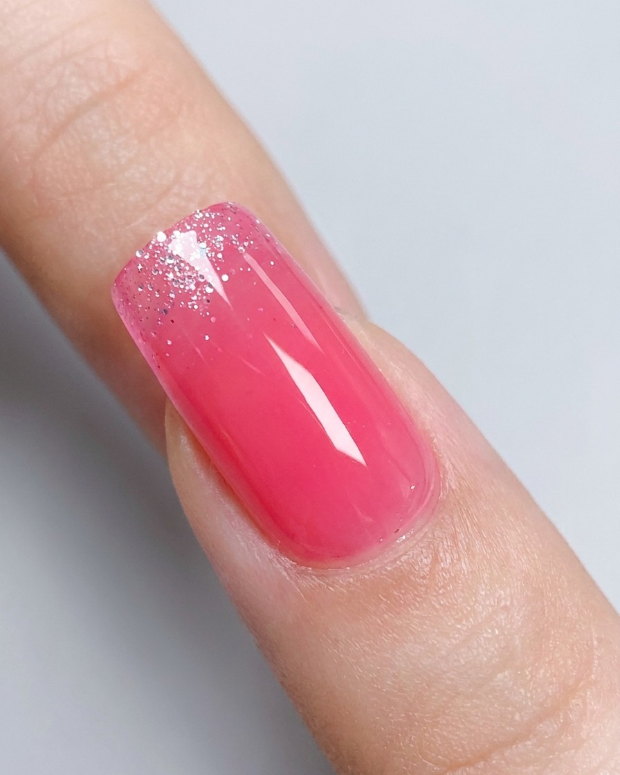 Un primo piano di ununghia sfumata in gel rosa lucido con un barattolo di gel glitter argento leggermente sfocato sullo sfondo.