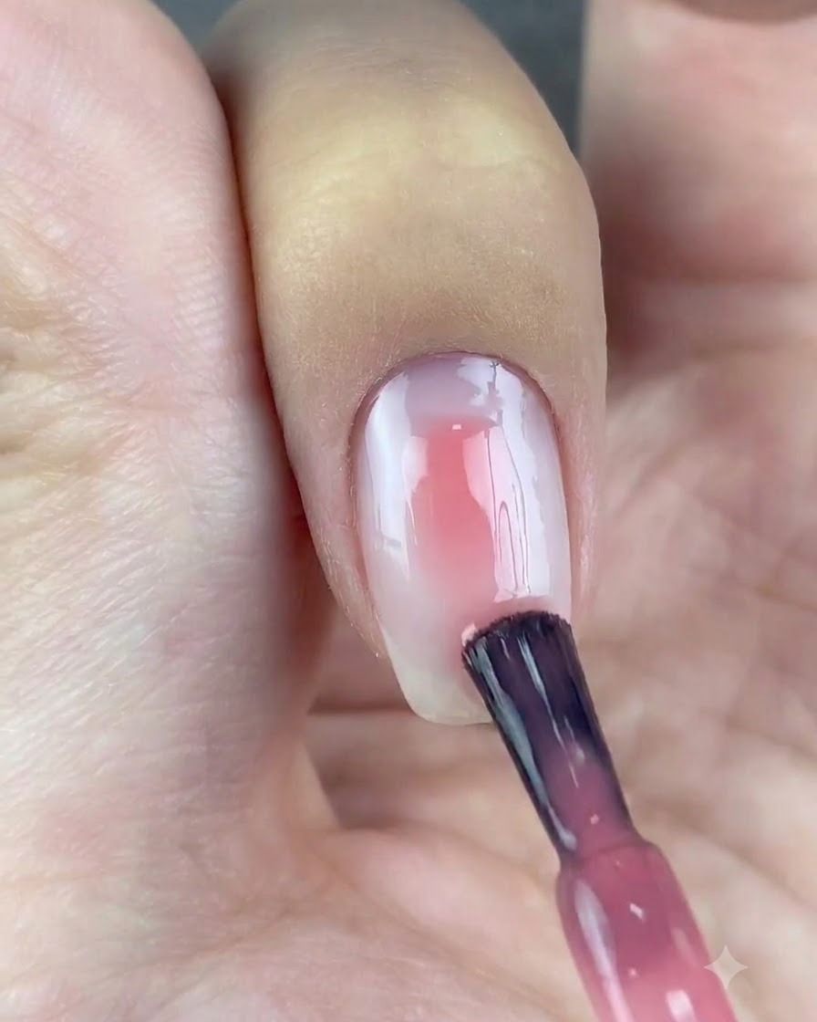 Un pennello per smalto applica una goccia di gel rosa traslucido al centro di ununghia con base bianca.
