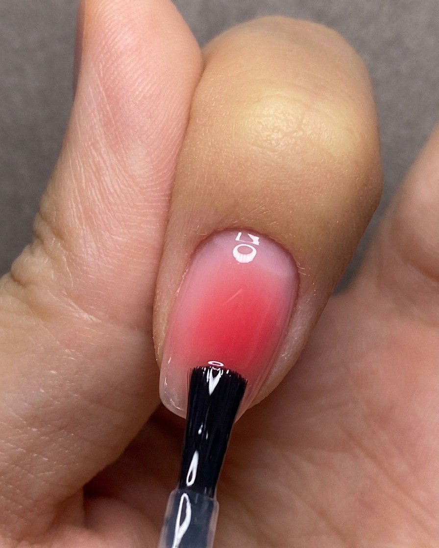 Un pennello applica un top coat trasparente in modo uniforme su ununghia con un design a sfumatura blush rossa.