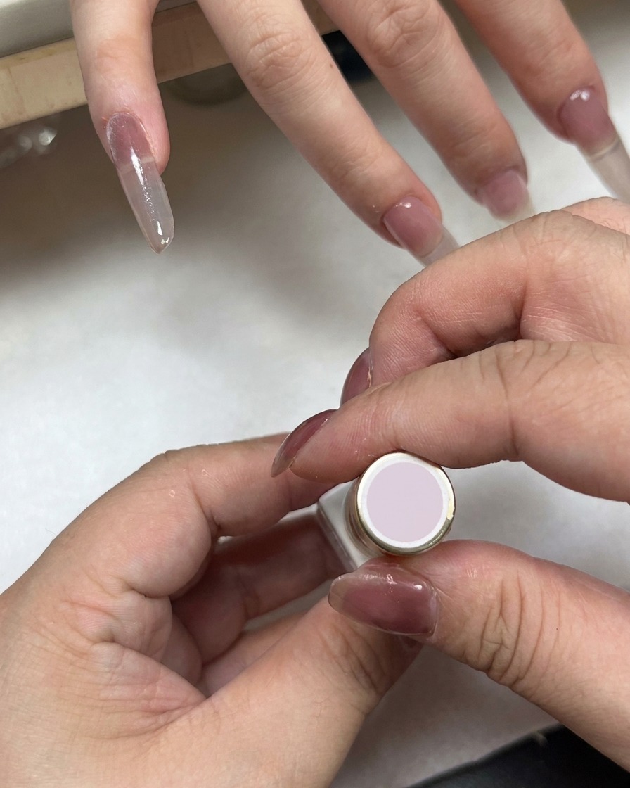 Un flacone di smalto con tappo rosa chiaro tenuto vicino a una mano con unghie sfumate.