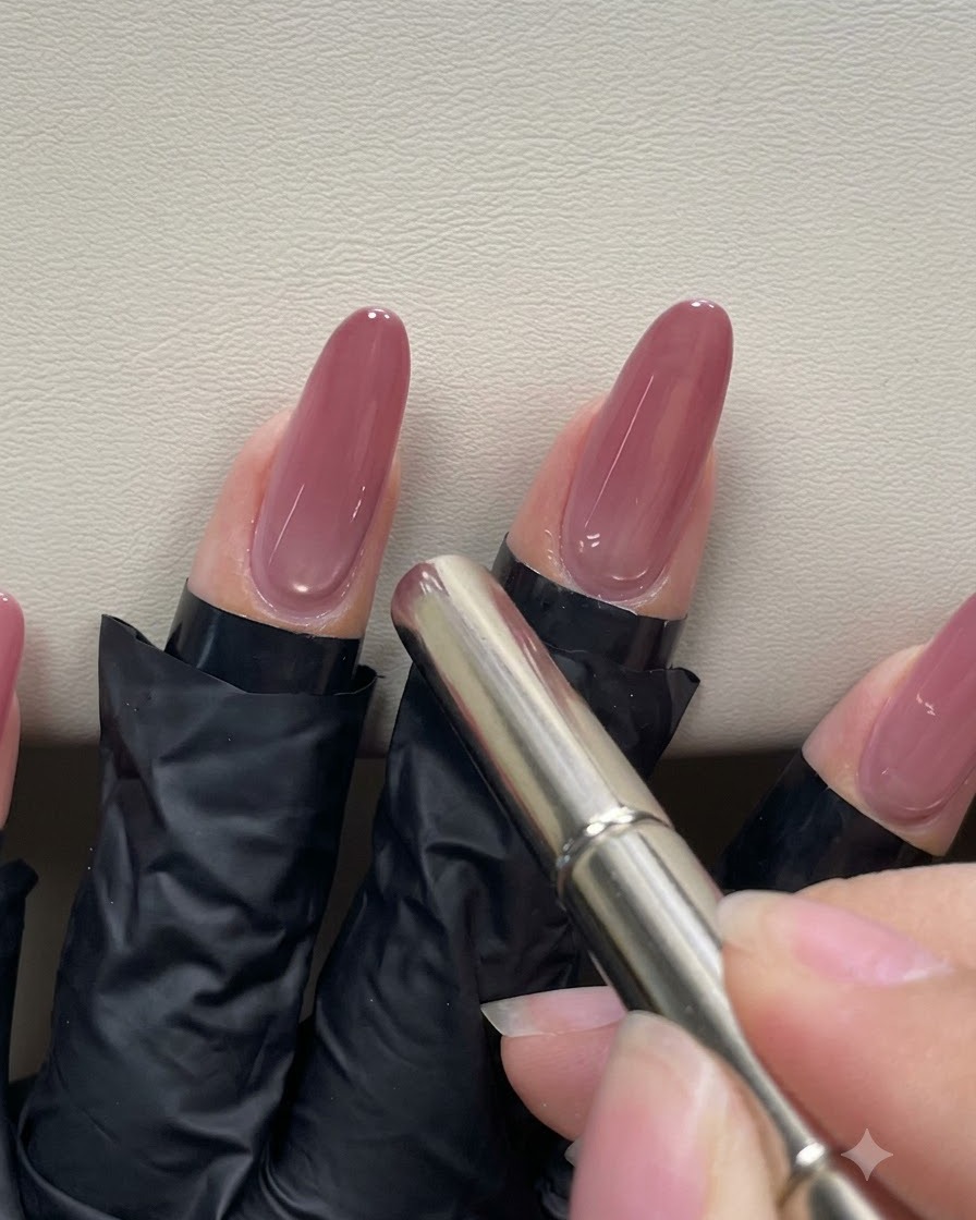 Una mano che usa un magnete cilindrico a penna vicino alle cuticole di ununghia in gel rosa pesca per manipolare il riflesso magnetico cat-eye.