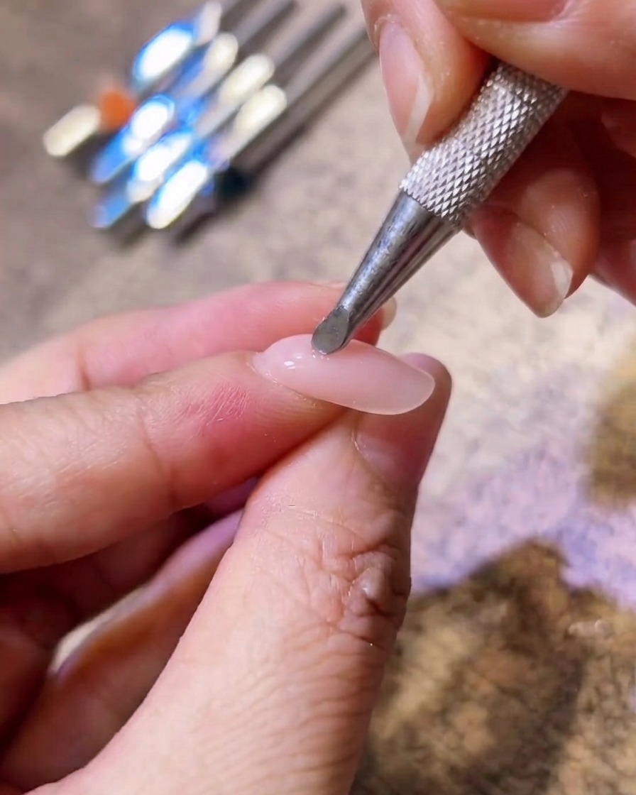 Primo piano di uno strumento applicatore in metallo che stende il pigmento rosa blush al centro di ununghia in gel nude.
