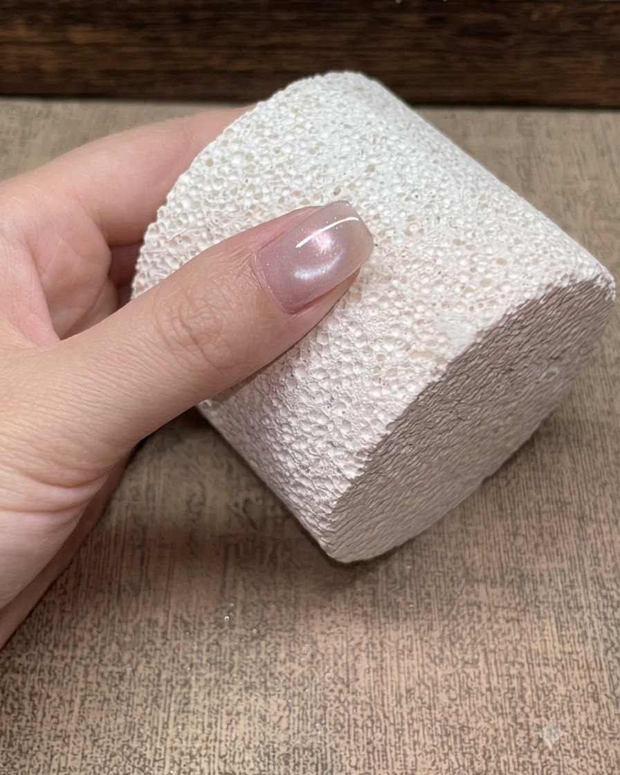 Primo piano di una singola unghia appoggiata su un blocco bianco testurizzato, che mostra un riflesso morbido cat-eye champagne effetto perla di vetro.