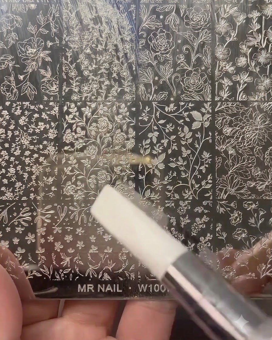 Un timbro per unghie trasparente che preme su una piastra di stampa in metallo incisa con vari motivi floreali e botanici.