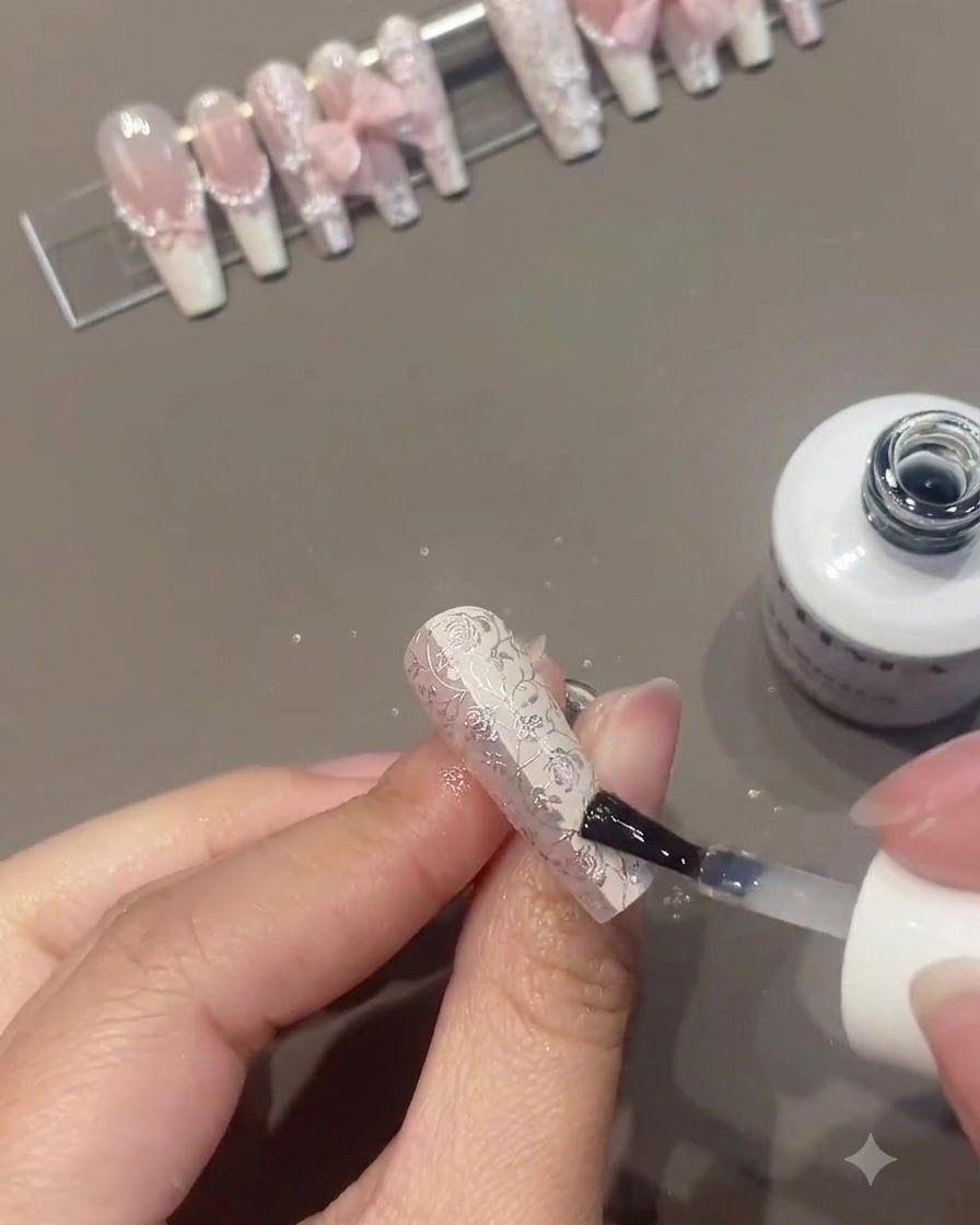 Un pennello per unghie che applica gel trasparente o top coat su ununghia floreale decorata in modo intricato mentre altre unghie finite sono esposte su un supporto sullo sfondo.