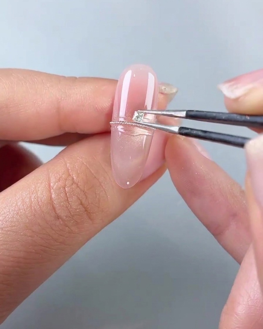 Una pinzetta posiziona un piccolo diamante quadrato accanto a una fila di perle dacciaio su ununghia in gel rosa.