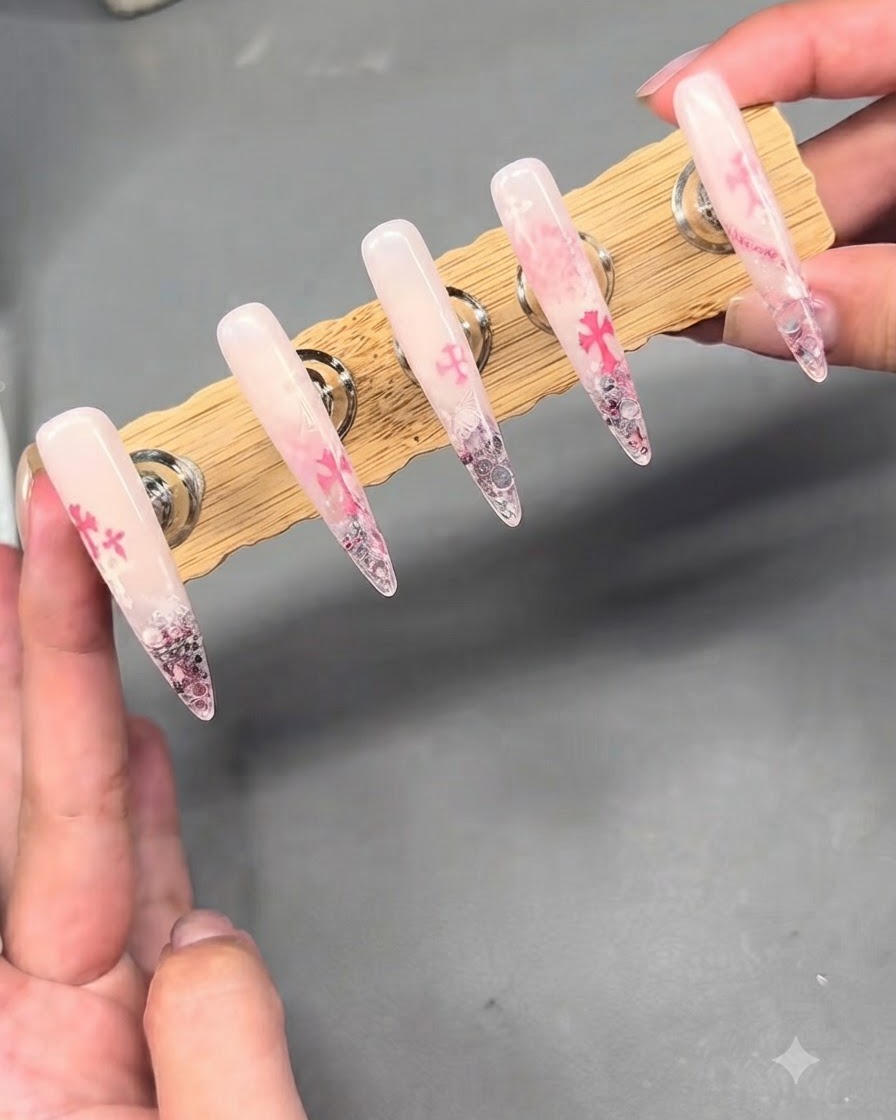 Un supporto di esposizione che contiene cinque lunghe unghie finte con sfumatura nude e rosa caratterizzate da stamping Chrome Hearts e abbellimenti in strass.
