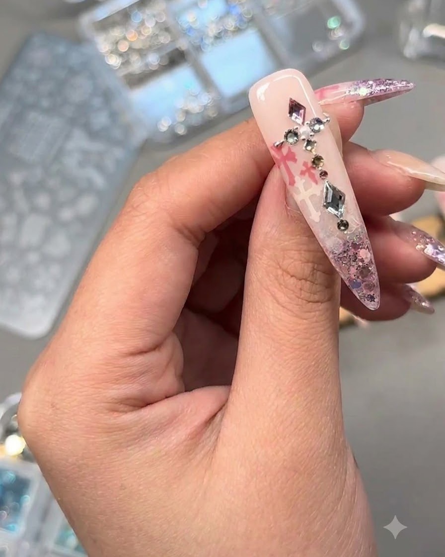 Primo piano di ununghia decorata adornata con cristalli, una perla e charm metallici su glitter rosa e arte stampata.