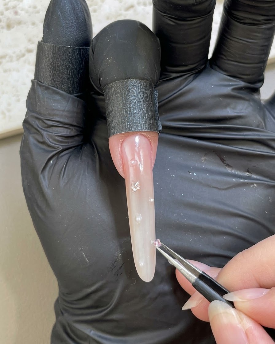 Una mano guantata che usa le pinzette per posizionare un piccolo strass rosa su una superficie ungueale decorata.