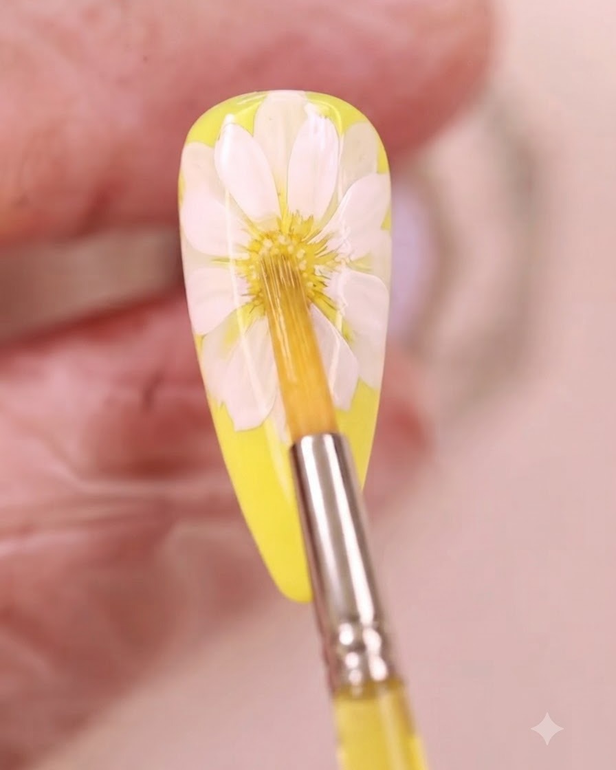 Un pennello sottile applica del liquido acquerello giallo al centro di un design di nail art con margherita bianca.
