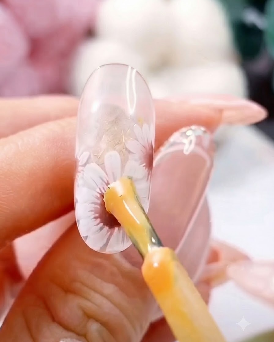 Un pennello applica un punto di gel giallo spesso al centro di una margherita bianca dipinta su ununghia trasparente.