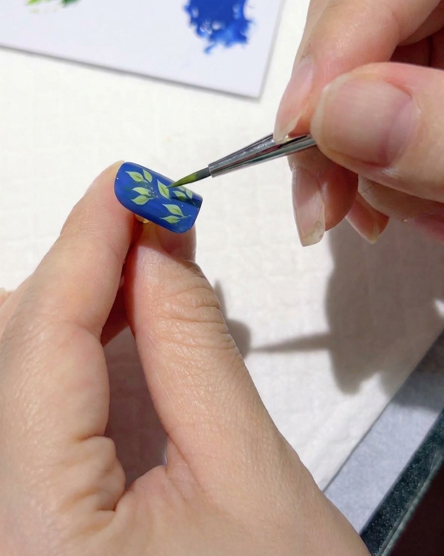 Un pennello per nail art fine che aggiunge tratti verde chiaro sopra le foglie scure su ununghia blu.
