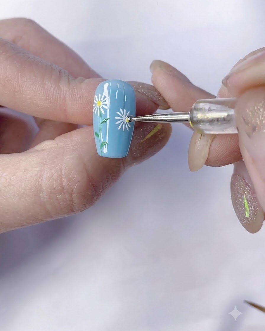 Un dotting tool applica un punto centrale giallo a una margherita bianca su ununghia blu.