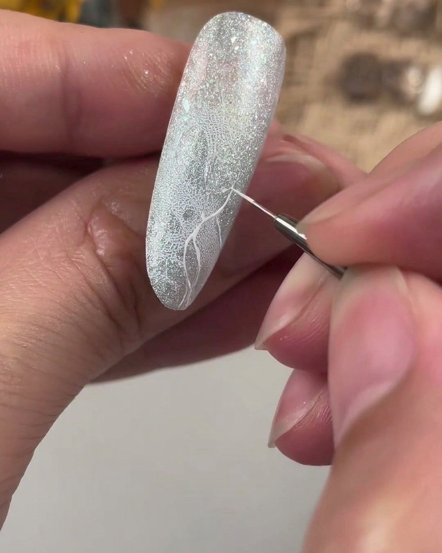 Un pennello da dettaglio fine che disegna una croce guida bianca su ununghia glitterata e testurizzata.