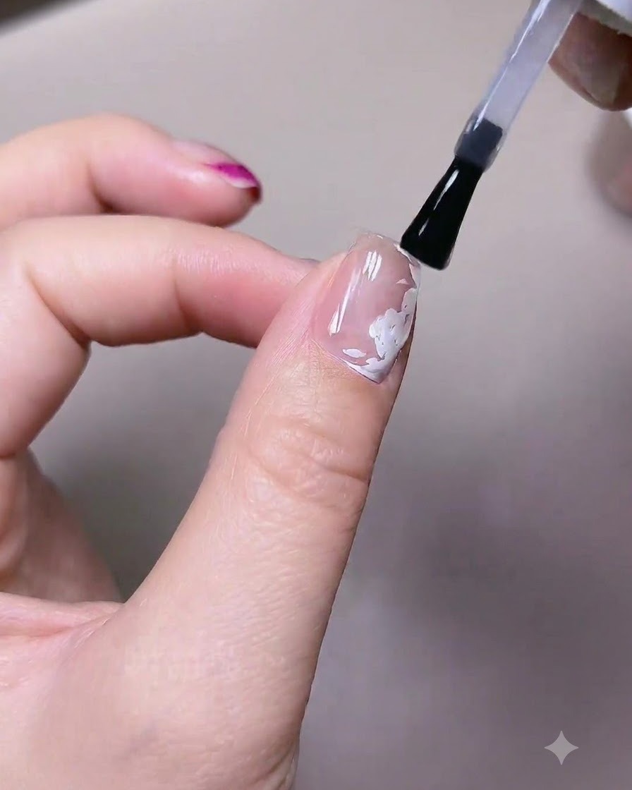 Un pennello applica uno strato di smalto top gel opaco su ununghia con elementi di rose bianche pre-dipinte.