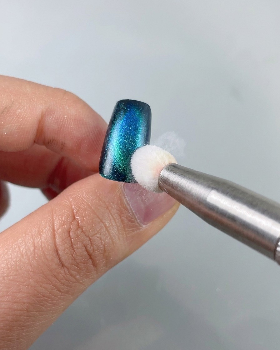 Una spugnetta tampona il gel da stamping nero sul bordo di unghia cat-eye verde-blu.